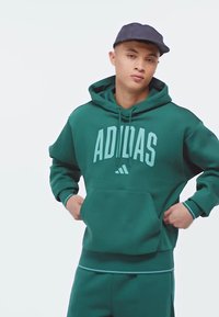 Haut à capuche en coton vert avec poche frontale et logo "ADIDAS". Doté d'une capuche avec cordon de serrage et de bords côtes, associé à un jogger assorti.