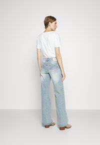 Light-wash denim jeans with a wide-leg cut, featuring red stitching and a vintage-style label at the back. Paired with a plain white t-shirt.