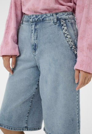 Gaiši zili džinsa šorti ar pīnumiem un krellēm rotātiem kabatām, valkāti kopā ar teksturētu rozā garās piedurknes topu.