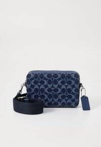 Tmavě modrá džínová kabelka přes rameno s kontrastním modrým vzorem loga, zapínán ím na zip, odnímatelným popruhem a malým detailním štítkem. Texturovaný materiál.