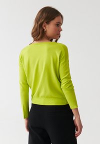 Limonengrüner Strickpullover mit langen Ärmeln und lockerer Passform, mit weitem Halsausschnitt und geripptem Saum. Kombiniert mit schwarzen Hosen.