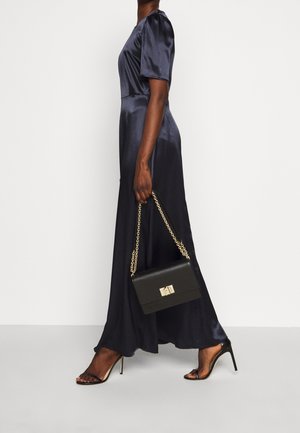 Femme en robe midi en satin bleu marine tenant un sac à main noir avec une bandoulière en chaîne dorée, portant des sandales à talons hauts noires sur un fond blanc.