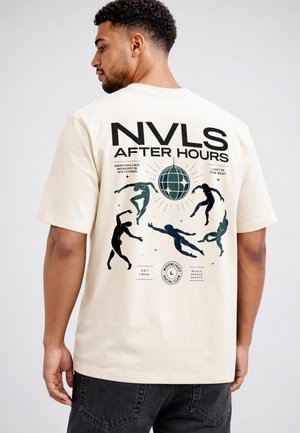 Man wearing beige shirt with black and green dancing figures, a disco ball, and text "NVLS After Hours" on the back.