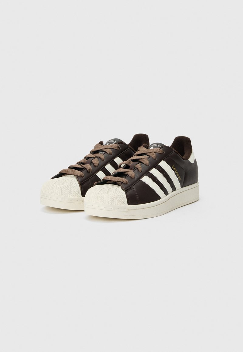 Coppia di sneakers Adidas Superstar color marrone scuro con punte a conchiglia bianche, tre strisce bianche, suole beige e lacci marroni su sfondo bianco.