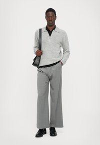 Suéter gris de punto con cremallera, polo negro debajo, pantalones anchos grises y zapatos negros, sosteniendo un bolso negro de hombro.