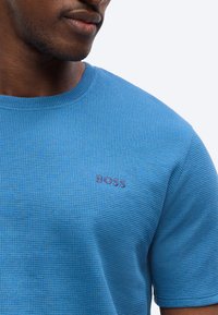 Κοντινό πλάνο ενός άνδρα που φοράει μπλε κοντομάνικο πουκάμισο με υφή, με το κέντημα «BOSS» στο αριστερό μέρος του στήθους.