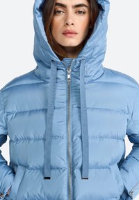 Ljusblå puffjacka med huva, med horisontella quiltade sektioner, framdragkedja och platta dragsnören. Slät, glänsande yta.