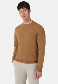 Maglione marrone lavorato a maglia con scollo rotondo e polsini e orlo a costine. Abbinato a pantaloni chiari e testurizzati.