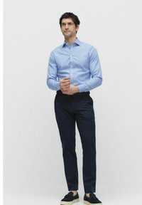 Lichtblauwe gestreepte overhemden met lange mouwen, gecombineerd met donkerblauwe broek en zwarte schoenen, staande tegen een effen achtergrond.