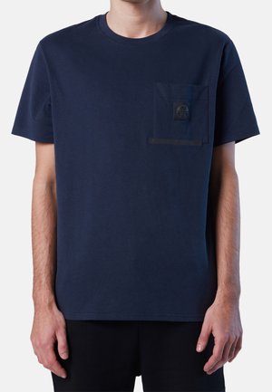 Mann trägt ein marineblaues T-Shirt mit kurzen Ärmeln und Brusttasche, auf der sich ein kleines rundes Logo-Patch befindet.