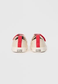 Baskets en toile beige avec un onglet de talon rouge et une doublure intérieure noire. Présente la marque "Converse All Star" sur la semelle en caoutchouc blanche.