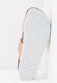Sandale avec semelle texturée ondulée blanche et brides en tissu beige, positionnée debout contre un arrière-plan d'angle blanc.