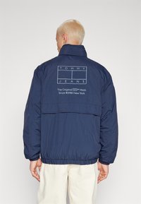 Tamsiai mėlyna pūkinė striukė su elastinga apatine dalimi ir rankogaliais, su balta atspauduota logo nugarinėje pusėje. Pagaminta iš lygaus, lengvo audinio.