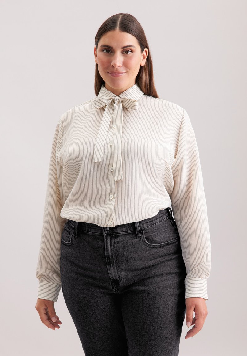 Blusa a righe crema con colletto a fiocco, chiusura con bottoni davanti, maniche lunghe, abbinata a jeans grigi scuri a vita alta.