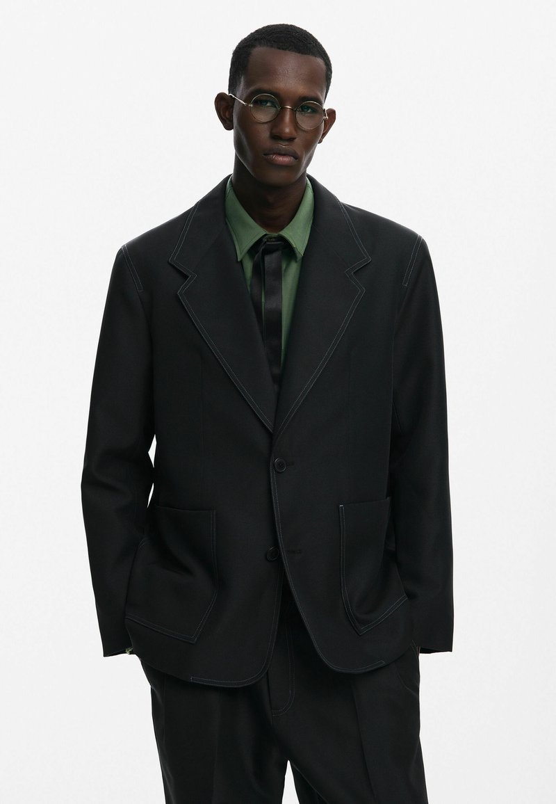 Fekete szabott zakó, kontrasztos fehér varrással, egy gombos záródással és két elülső zsebbel, zöld ing és fekete nyakkendő fölött viselve.