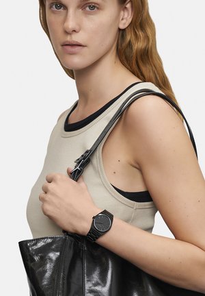 Svart armbandsur med en minimalistisk rund urtavla, svart armband. Ljusbeige linne med mörk kant, håller en blanka svart tote-bag.