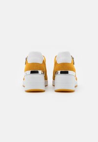 Żółte zamszowe sneakersy z białym, wyściełanym kołnierzem i błyszczącym srebrnym akcentem na pięcie. Podeszwa jest biała z pomarańczowym dnem.