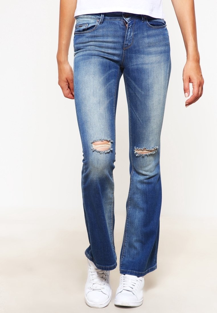 Personne portant un jean bleu déchiré aux genoux et des baskets blanches, debout devant un fond uni.