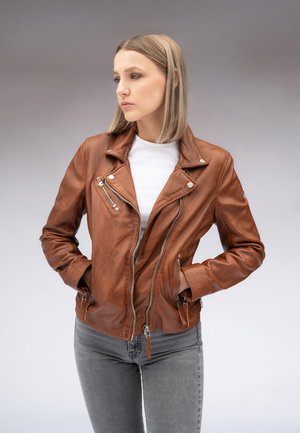Femme aux cheveux blonds raides portant une veste en cuir marron, une chemise blanche et un jean gris, debout les mains dans les poches de la veste.