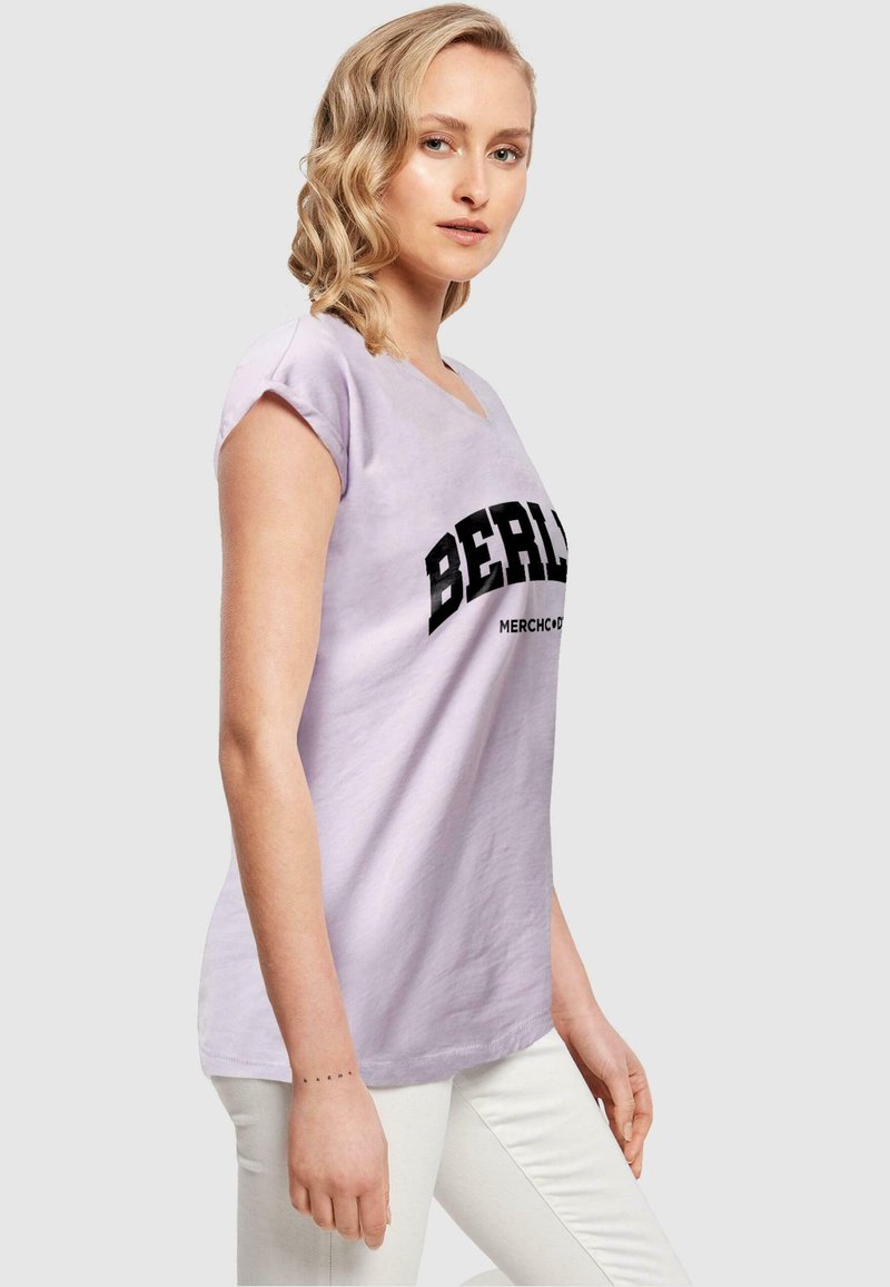Lyse lilla t-shirt med korte ærmer, der har teksten "BERLIN" i fed sort skrift og "MERCHANDISE" nedenunder, lavet af blødt bomuldsmateriale.
