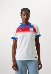 Balta Nike virsjakas ar sarkanu gradāciju un ziliem akcentiem. Iekļauj ribotu audumu, apaļu apkakli, īsām piedurknēm un Anglijas ģerboni.