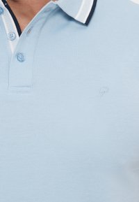 Ανοιχτό μπλε πόλο με σκούρο και λευκό ριγέ γιακά, τρία μπλε κουμπιά και μικρό κεντημένο γράμμα "P" στο στήθος.