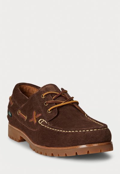 Zapato de embarcación de ante marrón con cordones marrones, costuras en contraste y suela de caucho. Presenta una etiqueta de logo en el lateral y detalles decorativos de cuero.