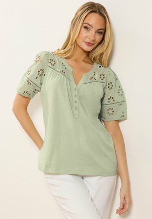 Vrouw met blond haar, gekleed in een lichtgroene blouse met bloemenuitsnijdingen op de mouwen en witte broek, staand tegen een lichte muur.