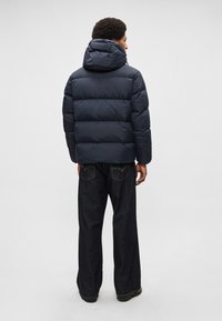 Casaco acolchoado azul-marinho com capuz, com seções acolchoadas para isolamento. Combinado com jeans escuros de corte largo.