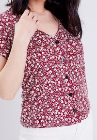 Blouse à imprimé floral rouge avec un décolleté en V, des manches courtes et des boutons noirs. Fabriquée dans un tissu léger avec des détails froncés.