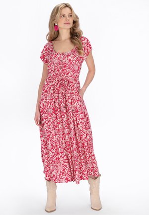 Hosszú szőke hajú nő piros-fehér virágos midi ruhában, rózsaszín fülbevalóval és bézs csizmával, egyik kezét a zsebében tartva áll.