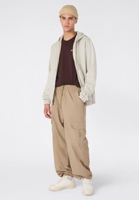 Jasnoszara bluza na zamek, ciemnobrązowa koszulka, beżowe spodnie cargo i białe trampki. Dodatkowe szczegóły to dzianinowa beżowa czapka.