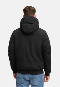 Man in een zwart kapvest en blauwe jeans, met zijn rug naar de camera tegen een effen witte achtergrond.