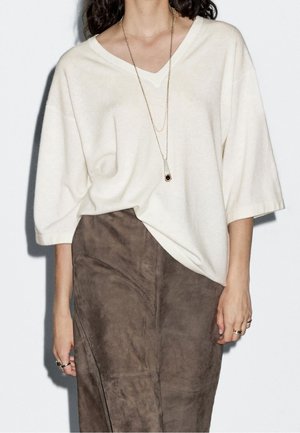 Kobieta w luźnej białej bluzce z dekoltem w serek, brązowej zamszowej spódnicy, długim złotym naszyjniku z wisiorkiem i pierścionkach na obu dłoniach na białym tle.