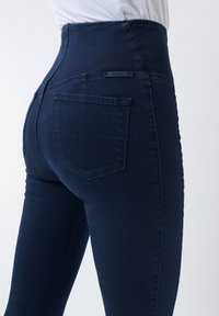 Tmavě modré džínové kalhoty s vysokým pasem a slim fit střihem, vybavené zadní kapsou a prošíváním kolem pasu.