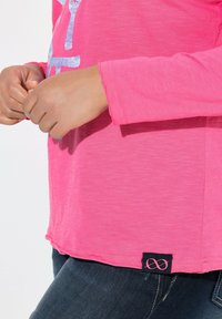 Långärmad, rosa T-shirt med en texturerad finish, med en grafisk design. Inkluderar en svart etikett med en oändlighetssymbol i nederkanten.