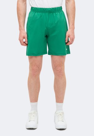 Άνδρας με φωτεινά πράσινα ελαστικά σορτς με λάστιχο στη μέση, λευκές κάλτσες και λευκά αθλητικά παπούτσια, στέκεται με τα χέρια χαλαρά δίπλα στο σώμα.