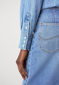 Detail světle modré denimové košile s knoflíkovým manžetou, obsahující čtyři krémové knoflíky a kapsu na boční straně oděvu.