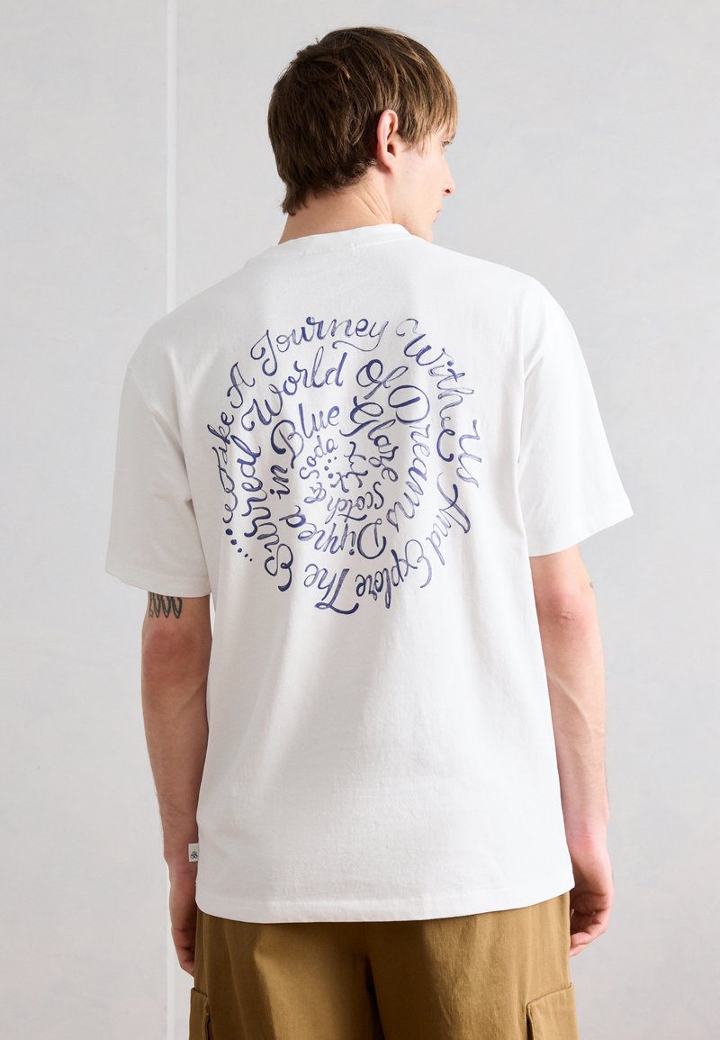 Άνδρας που φοράει λευκό T-shirt με σπειροειδές μπλε χειρόγραφο κείμενο στην πλάτη και καφέ cargo σορτς μπροστά σε απλό φόντο.