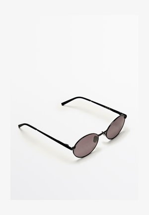 Vrouw met ronde zwarte zonnebril en een mouwloos bruin-zwart gebreid topje tegen een effen lichtgrijze achtergrond.