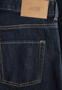 Jean en denim bleu foncé avec des coutures jaunes, montrant une poche arrière et une étiquette en cuir beige avec les lettres JJXX.
