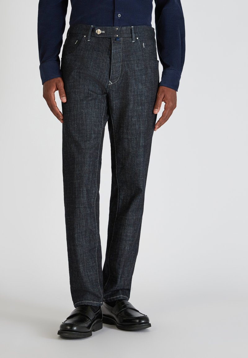 Mann in dunkelblauen geraden Jeans, schwarzen Lederloafers und einem navyblauen Langarmshirt, der vor einem schlichten Hintergrund steht.