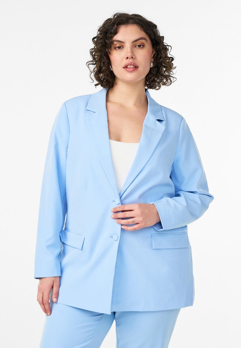 Frau mit lockigem Haar, die eine hellblaue Anzugjacke und passende Hose trägt, posiert mit einer Hand, die den Vorderteil der Jacke richtet.