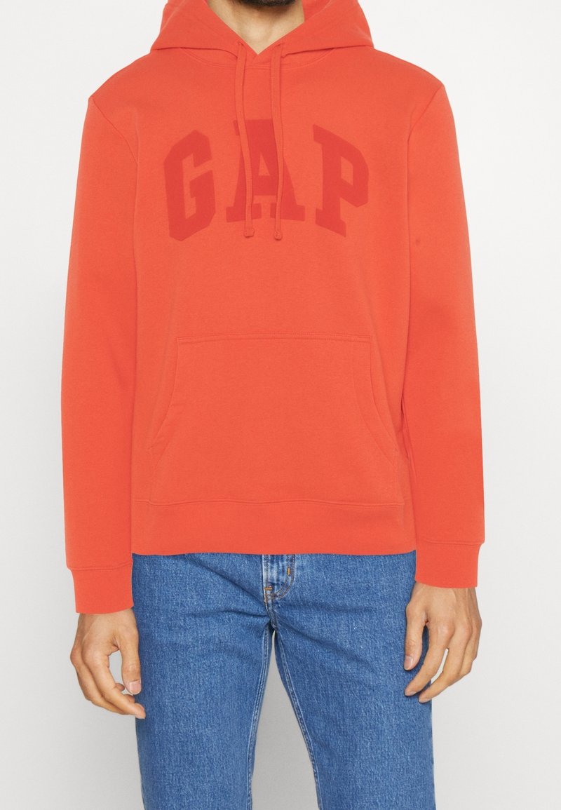 Homme portant un sweat à capuche orange GAP avec une poche avant et un jean bleu, présenté du cou aux hanches sur un fond clair.