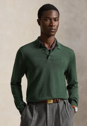 Homme portant un polo à manches longues vert foncé, un pantalon plissé gris, une écharpe à motif, une ceinture marron et une montre-bracelet sur un fond uni.