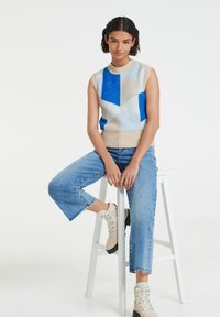 Woman with short dark hair sits on white stool wearing sleeveless geometric blue and beige sweater, cropped blue jeans, and beige boots.