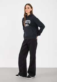 Zwarte hoodie met de print "Tokyo 1963", gecombineerd met wijdvallende zwarte jeans en donkere sneakers met witte zolen, tegen een effen achtergrond.