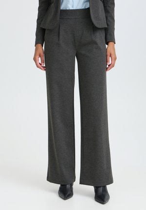 Vrouw die donkergrijze wijde broek, zwarte puntige laarzen, een lichtblauw overhemd en een donkergrijze blazer draagt.