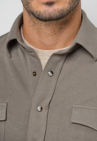 Chemise grise boutonnée avec un col, comprenant deux poches poitrine et des boutons décoratifs. Confectionnée en tissu doux avec une texture lisse.