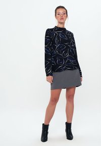Maglione nero con motivo a foglie multicolore, maniche lunghe, abbinato a una gonna grigia. Modella indossa stivaletti neri alla caviglia. Sfondo semplice.